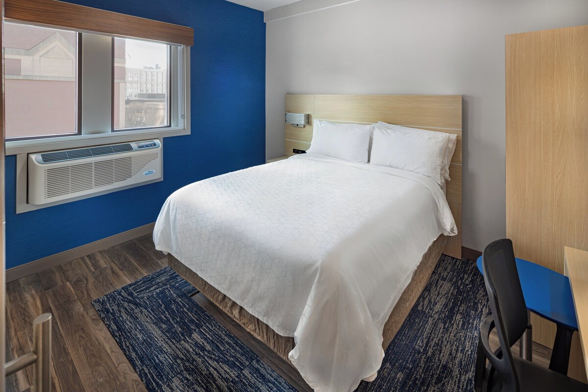 A hotel room with a bed, a desk, a chair, a window, and a wall-mounted air conditioner.