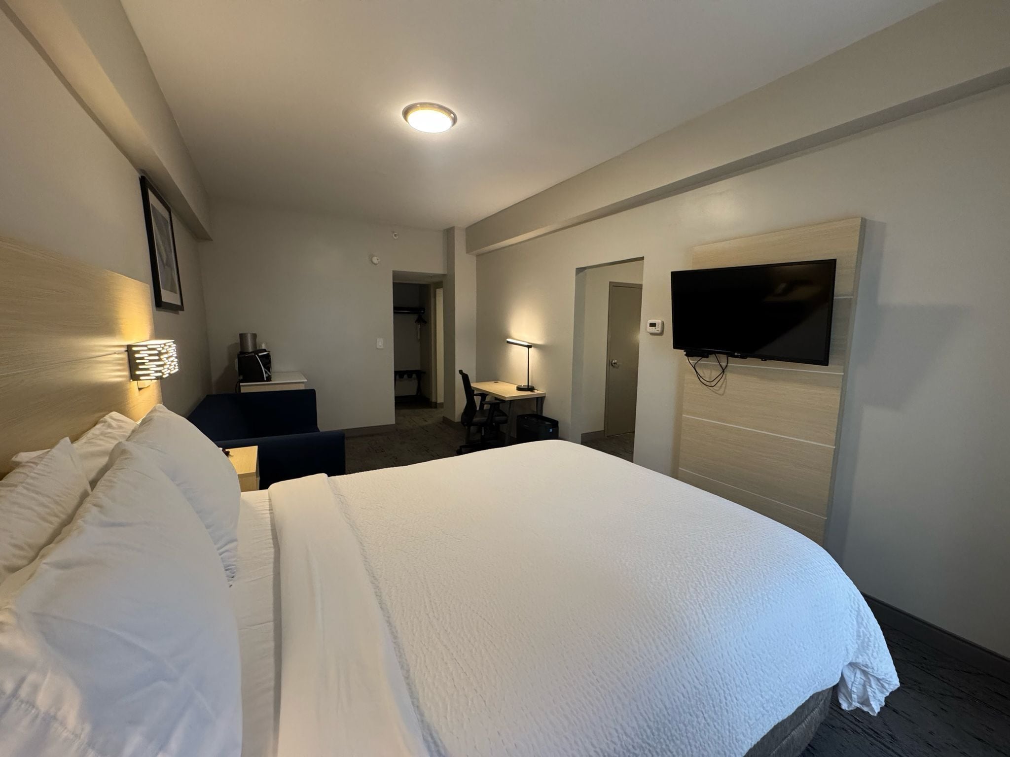 A hotel room with a large bed, a desk, a chair, a TV mounted on the wall, and a door leading to another room.