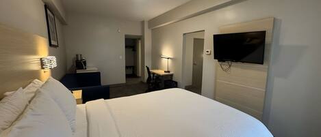 A hotel room with a large bed, a desk, a chair, a TV mounted on the wall, and a door leading to another room.