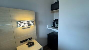 A modern hotel room with a wooden panel wall, a bedside table with a digital clock, a coffee machine, and a small black appliance.