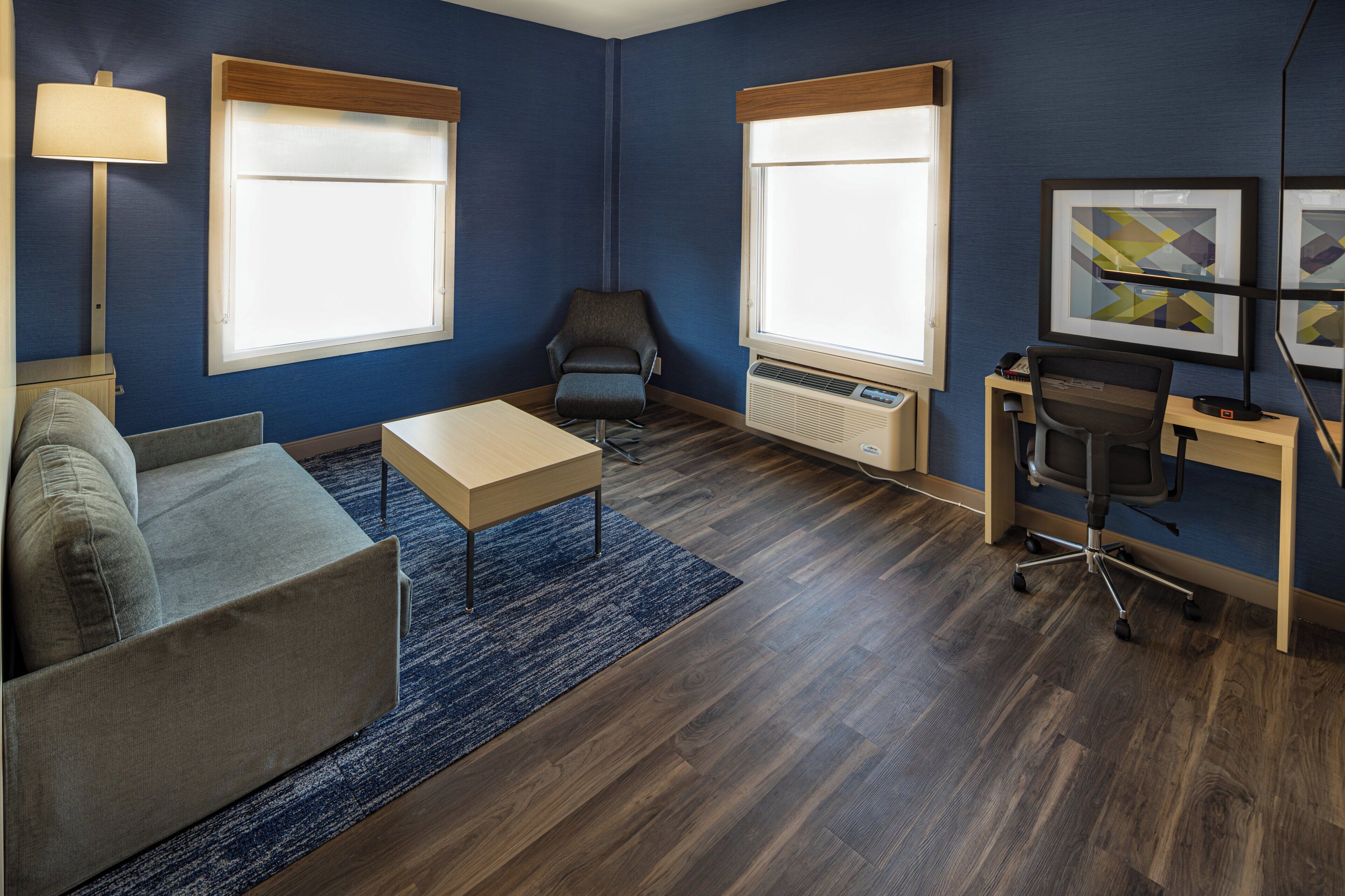 Une chambre d’hôtel moderne équipée d’un canapé, d’une petite table, d’un bureau avec une chaise et de deux fenêtres avec des stores.