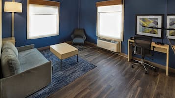 A modern hotel room with a sofa, a small table, a desk with a chair, and two windows with blinds.