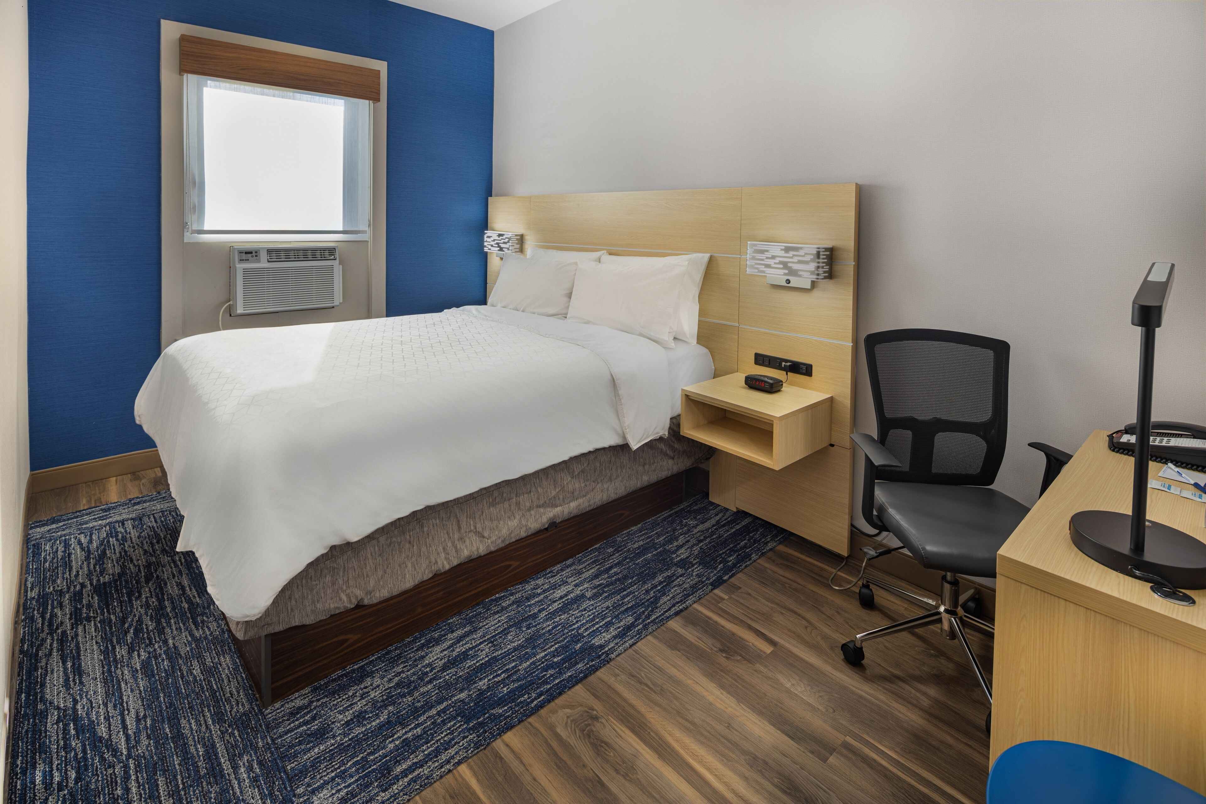 A hotel room with a bed, a desk, a chair, a lamp, and a window.