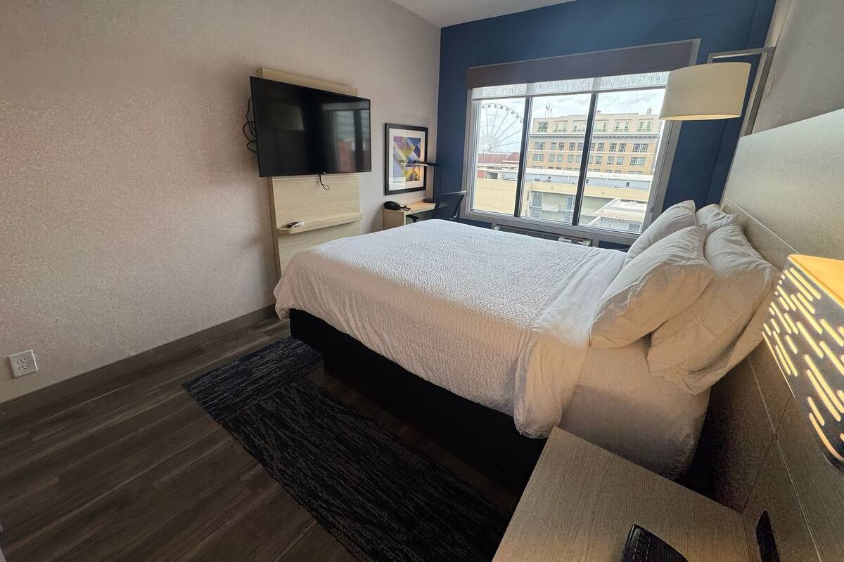 A bedroom with a bed, a TV mounted on the wall, a window with a view of buildings, and a lamp on the nightstand.