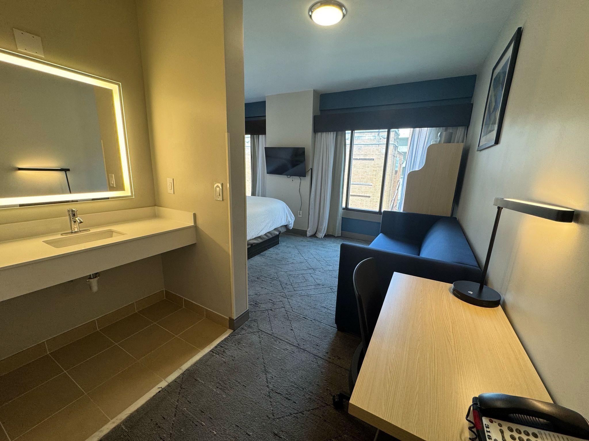 A hotel room with a large mirror, a sink, a bed, a desk, and a chair.