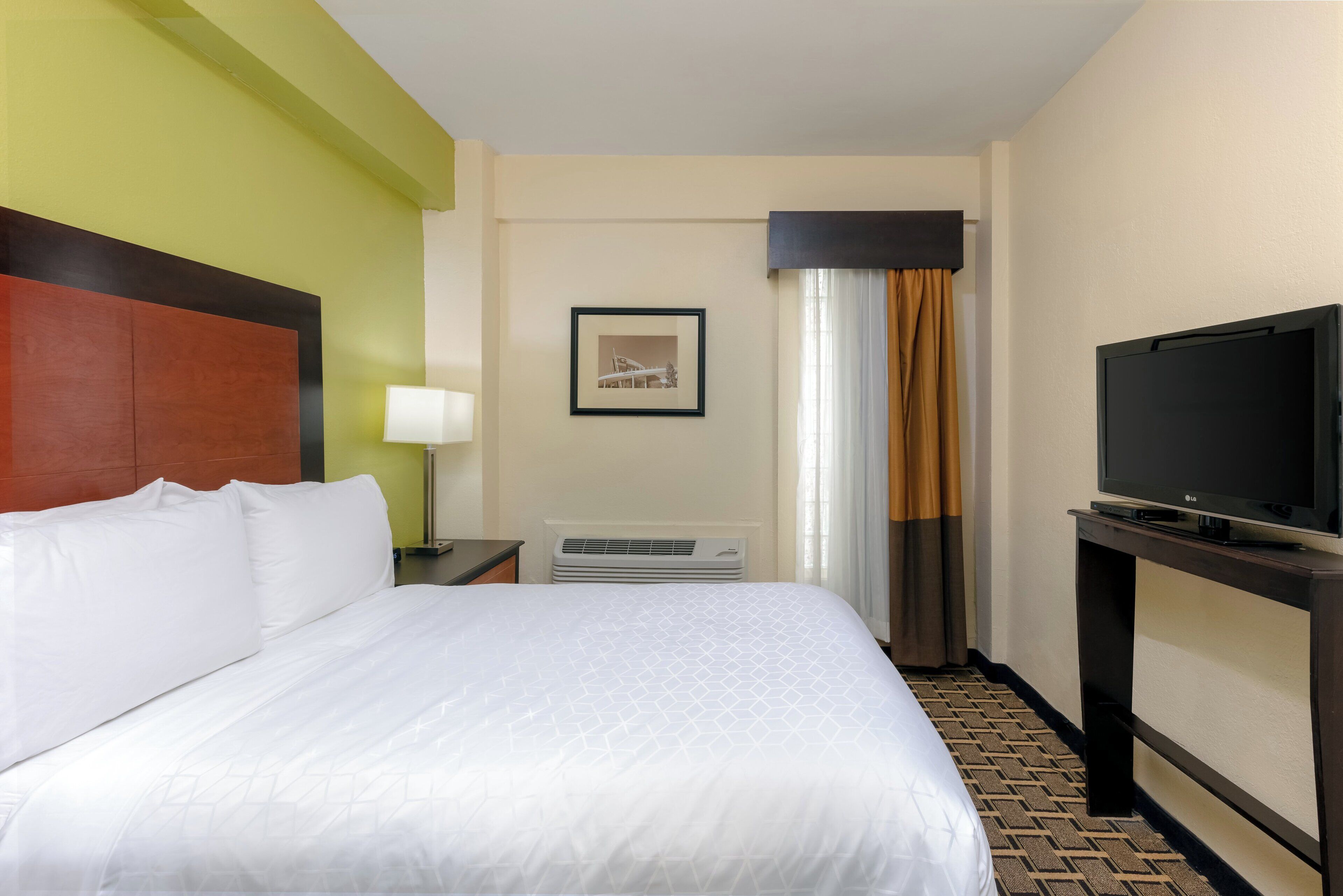 A hotel room with a bed, a television, a framed picture on the wall, and a window with curtains.