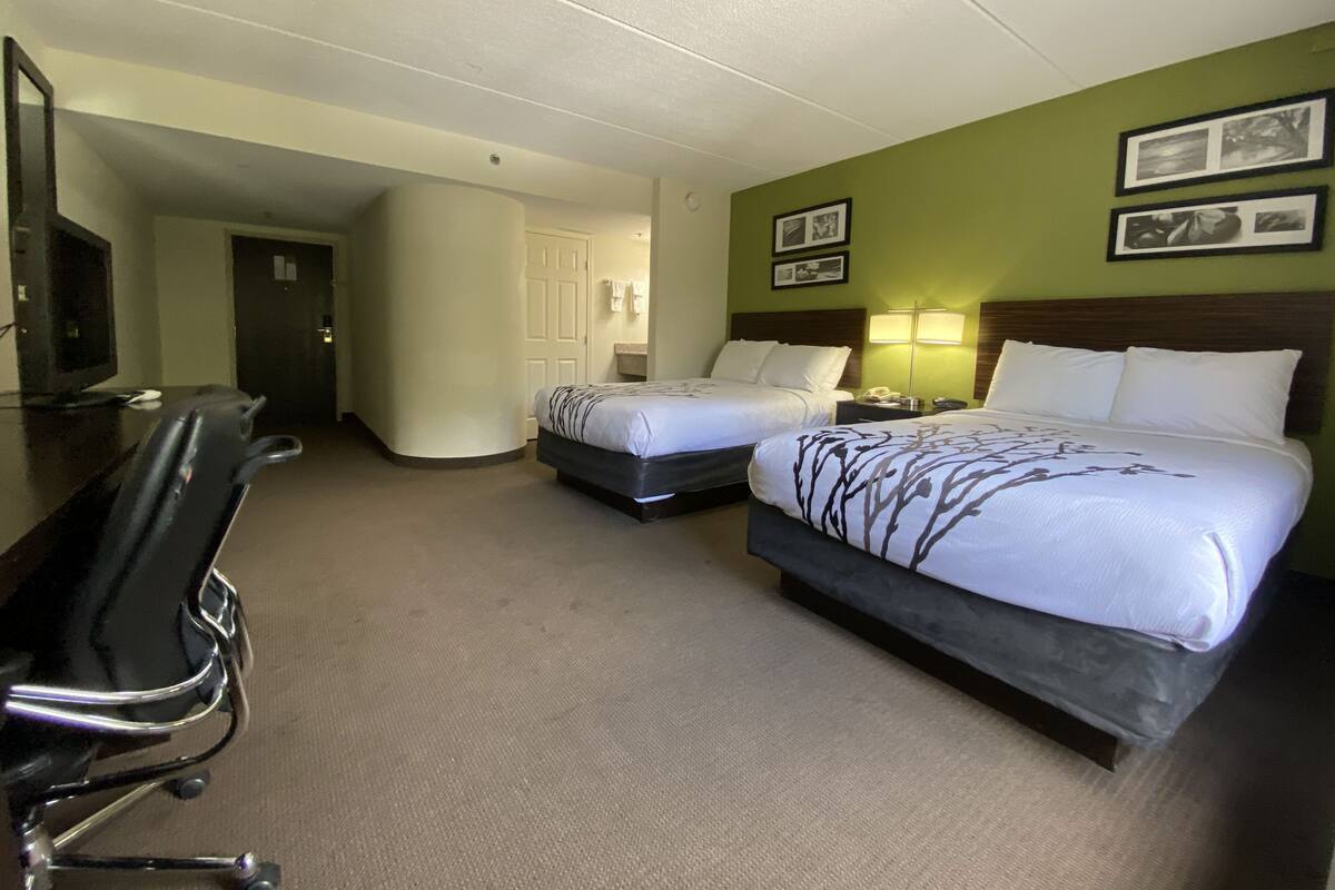 A hotel room with two beds, a desk, and a chair. There are two lamps on the nightstands, a television on the desk, and a door leading to another room.