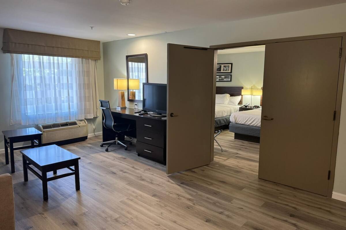 Ein Hotelzimmer mit Schreibtisch, Stuhl und Bett. Ein Fenster mit Vorhängen, eine Lampe und ein Spiegel sind an der Wand angebracht.
