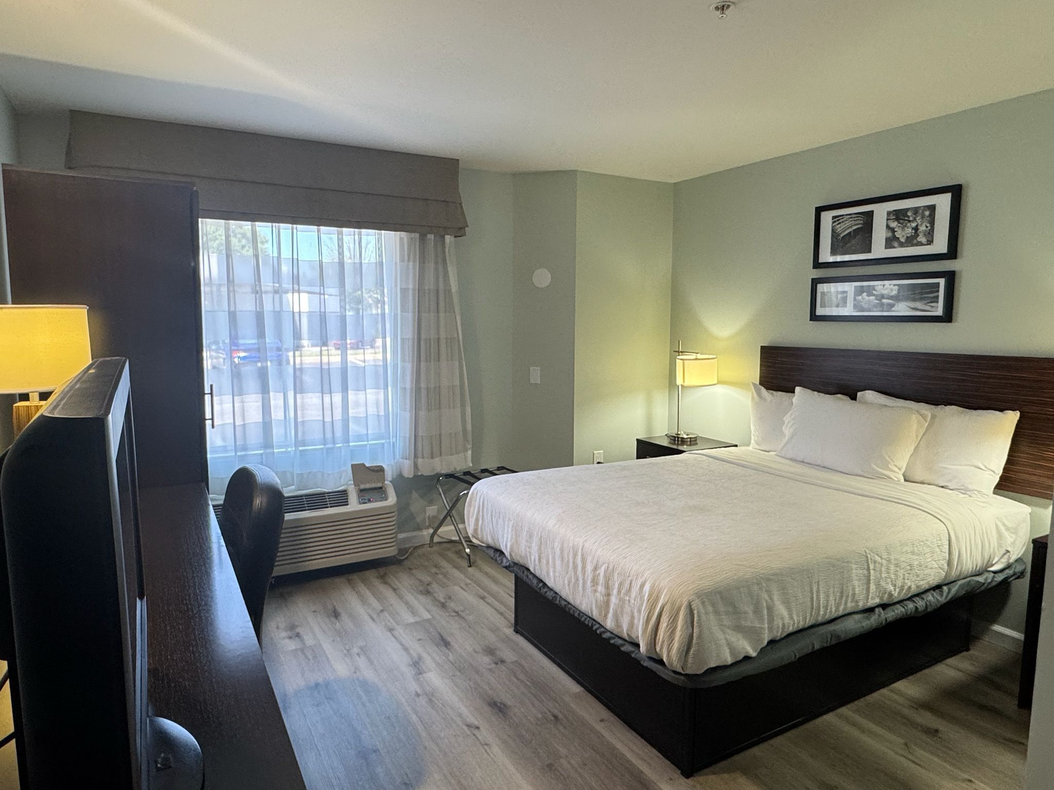A hotel room with a bed, a TV, a window with curtains, a wall-mounted lamp, and two framed pictures.