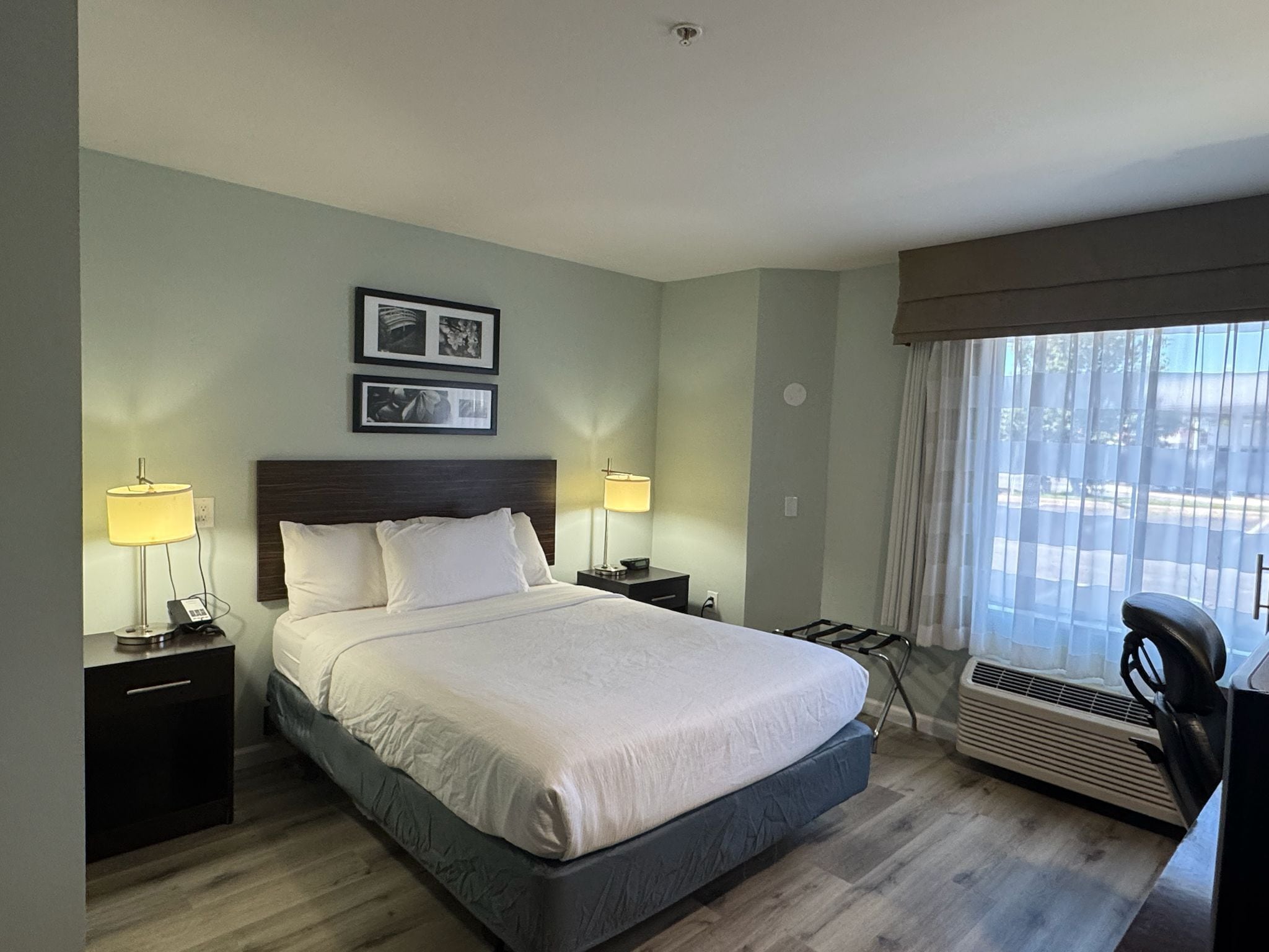 Habitación de hotel con una cama, dos lámparas de noche, un calefactor, un escritorio con una silla y dos cuadros en la pared.