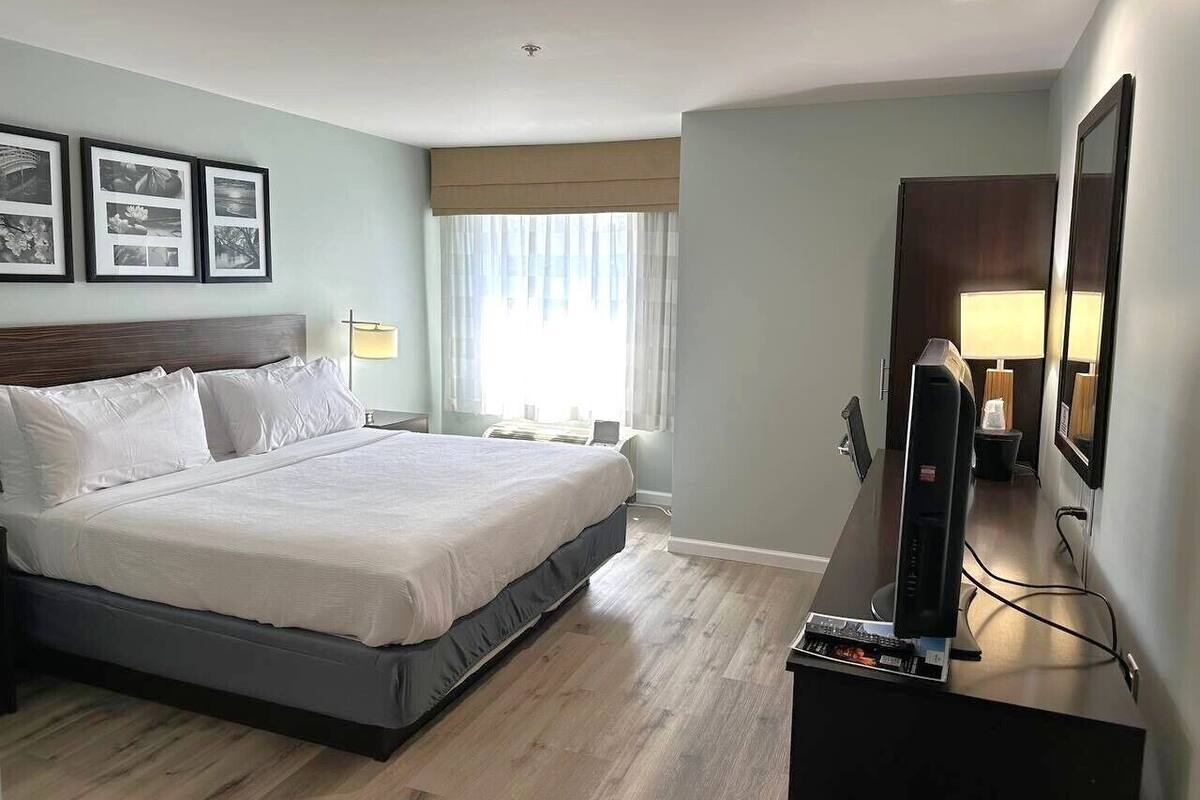 Ein Hotelzimmer mit einem großen Bett, einem Flachbildfernseher, einem Schreibtisch mit Computer und einem Fenster mit Vorhängen.