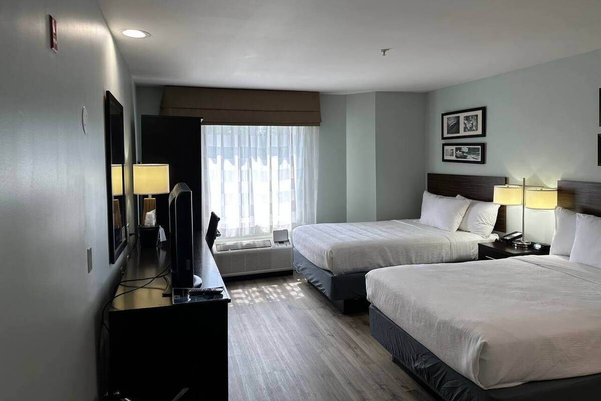 Ein Hotelzimmer mit zwei Betten, einem Schreibtisch mit Computer, einem Fernseher und einem Fenster mit Vorhängen.