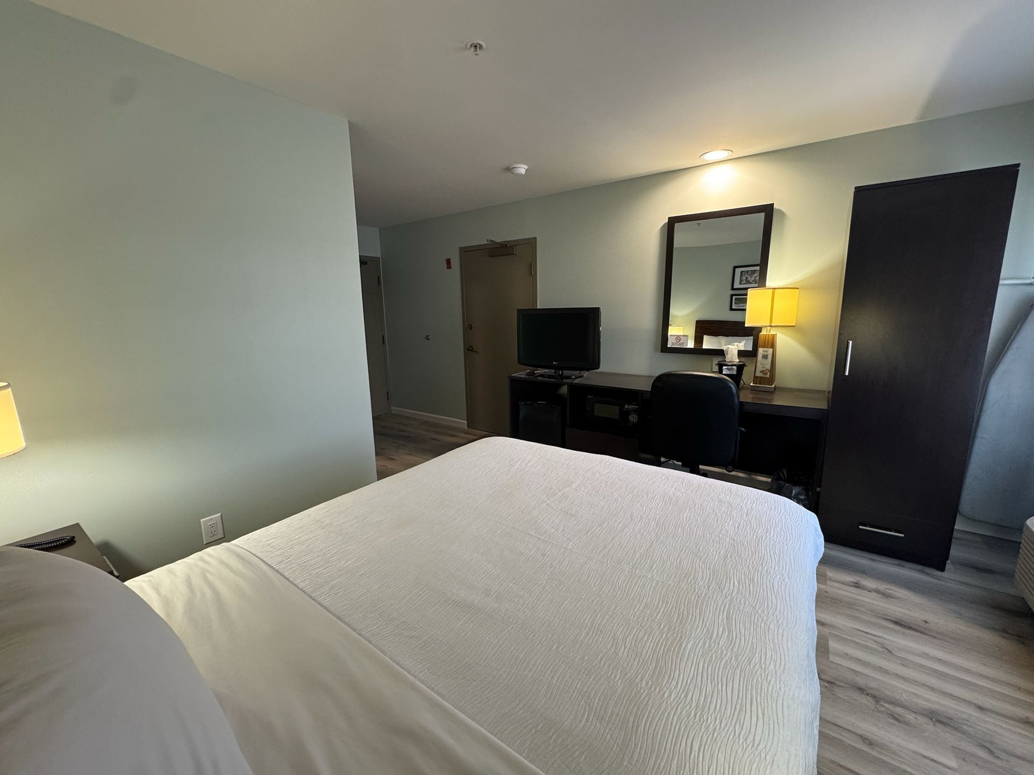 Una habitación de hotel con una cama, un escritorio con computadora, un televisor, un espejo y un armario.