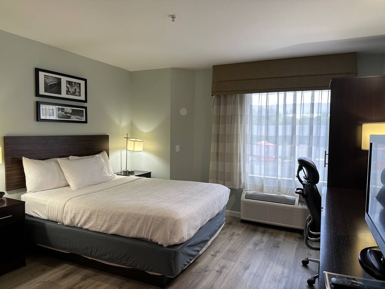 A hotel room with a bed, bedside table, lamp, air conditioning unit, and a television.