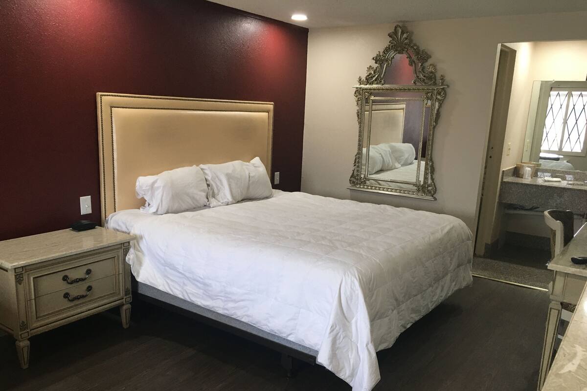 A large bed with a headboard, a bedside table with drawers, a mirror, and a bathroom visible in the background.