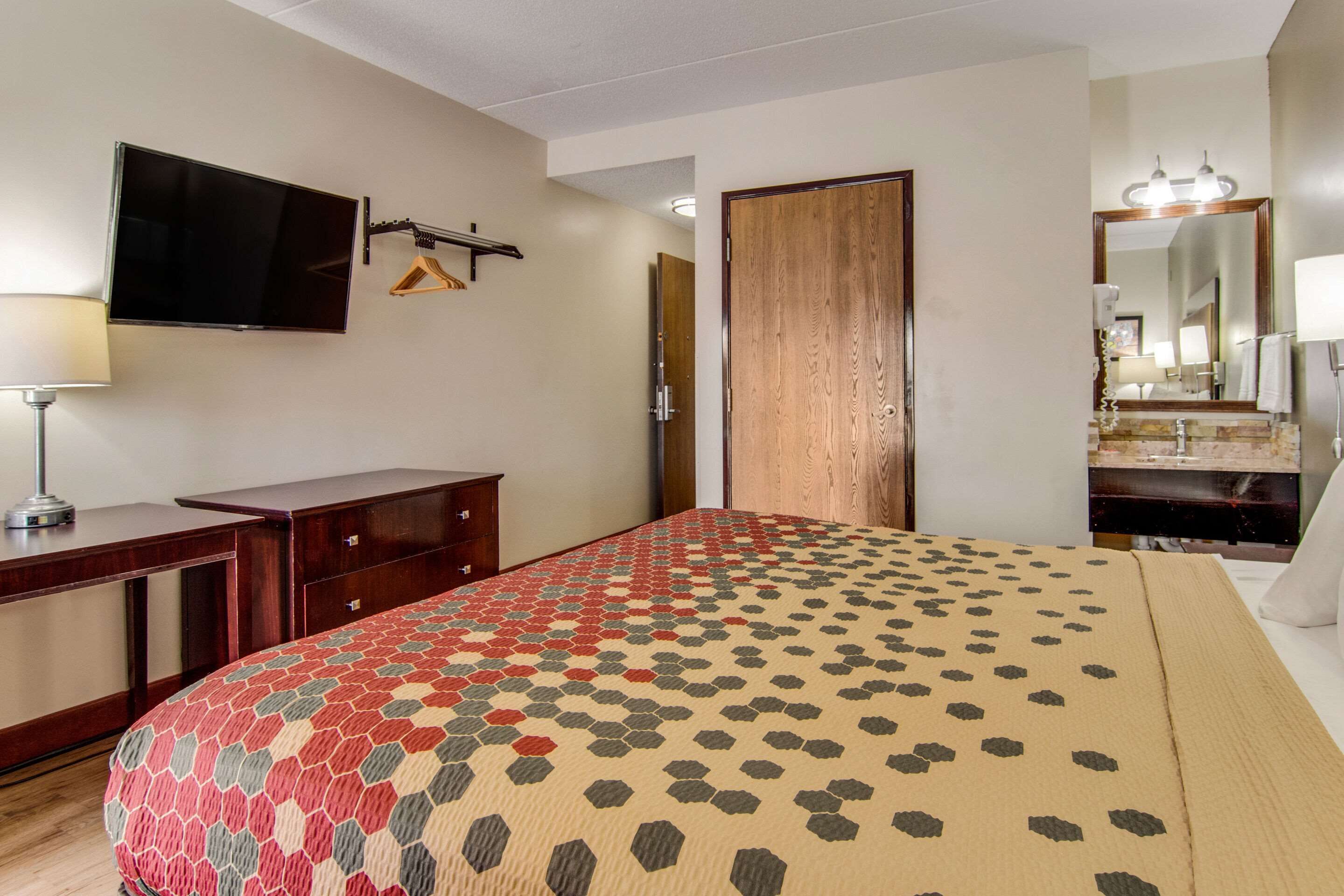 Una habitación de hotel con cama, televisor montado en la pared, armario de madera, espejo con iluminación y baño con lavamanos y espejo.