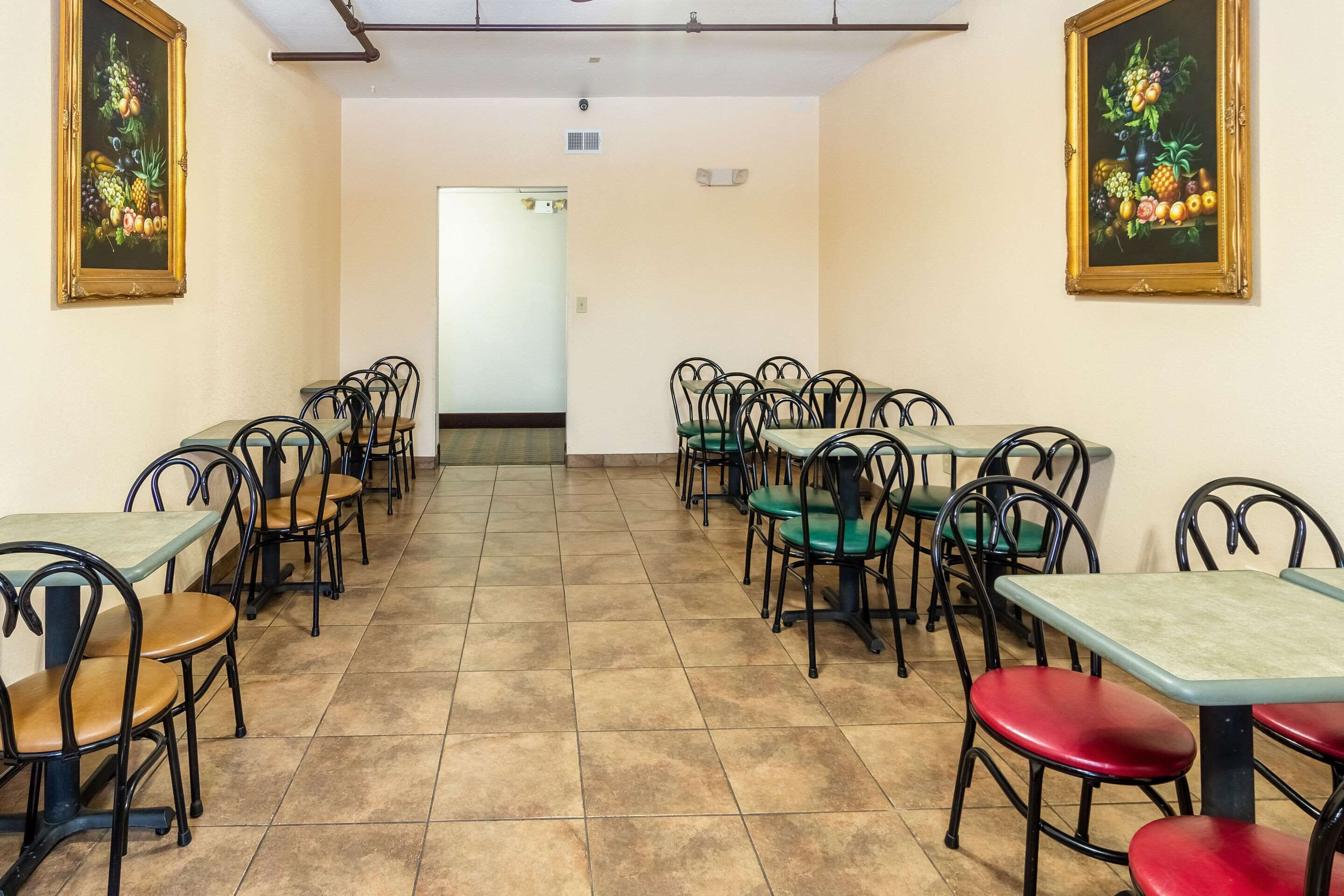 Un'area pranzo con tavoli e sedie disposti in file, un pavimento piastrellato e due quadri appesi alle pareti.