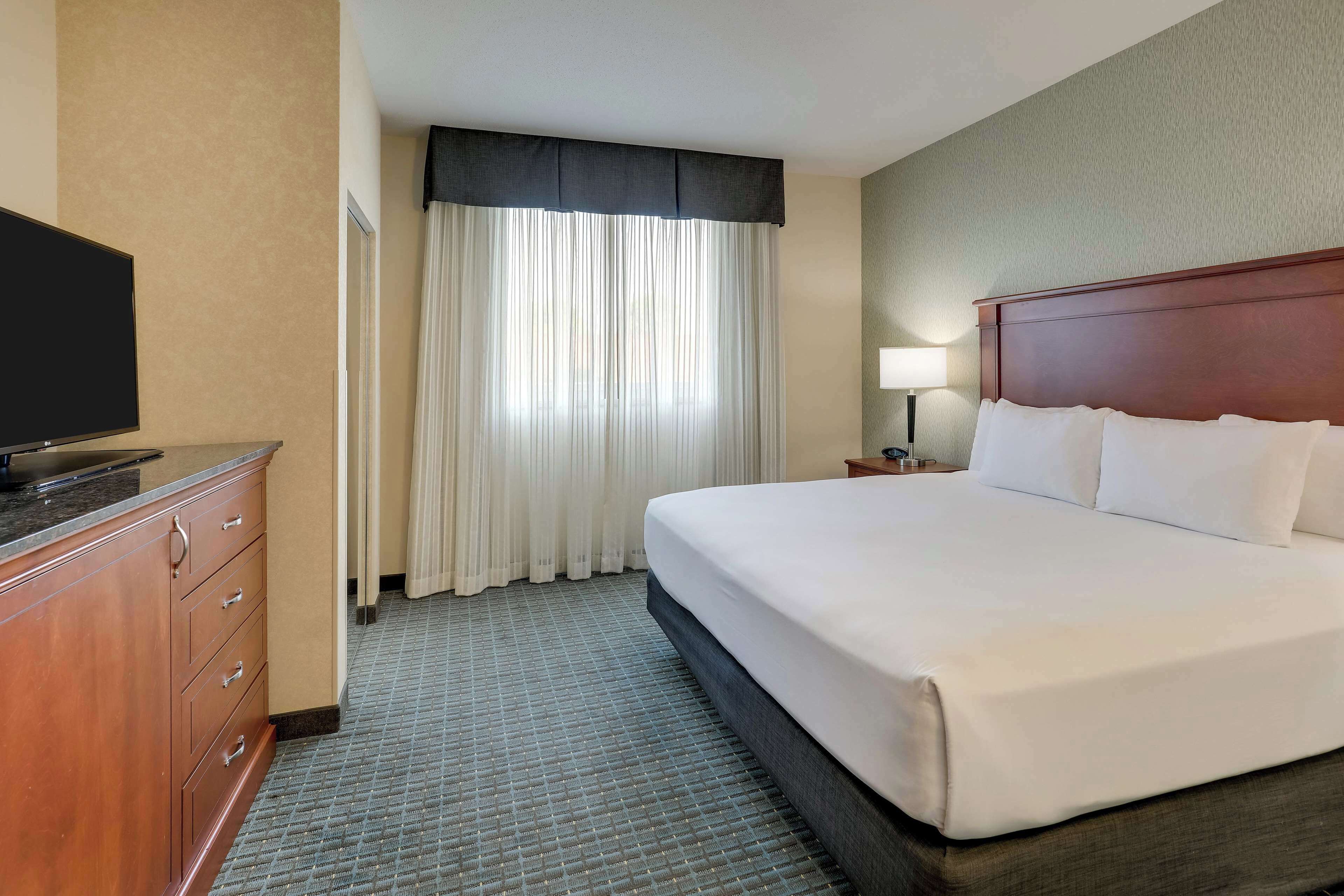 A hotel room with a large bed, a TV on a wooden cabinet, a nightstand with a lamp, and a window with curtains.