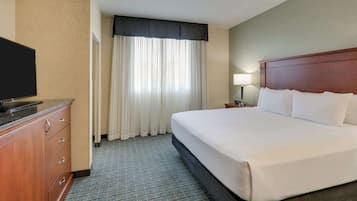 A hotel room with a large bed, a TV on a wooden cabinet, a nightstand with a lamp, and a window with curtains.