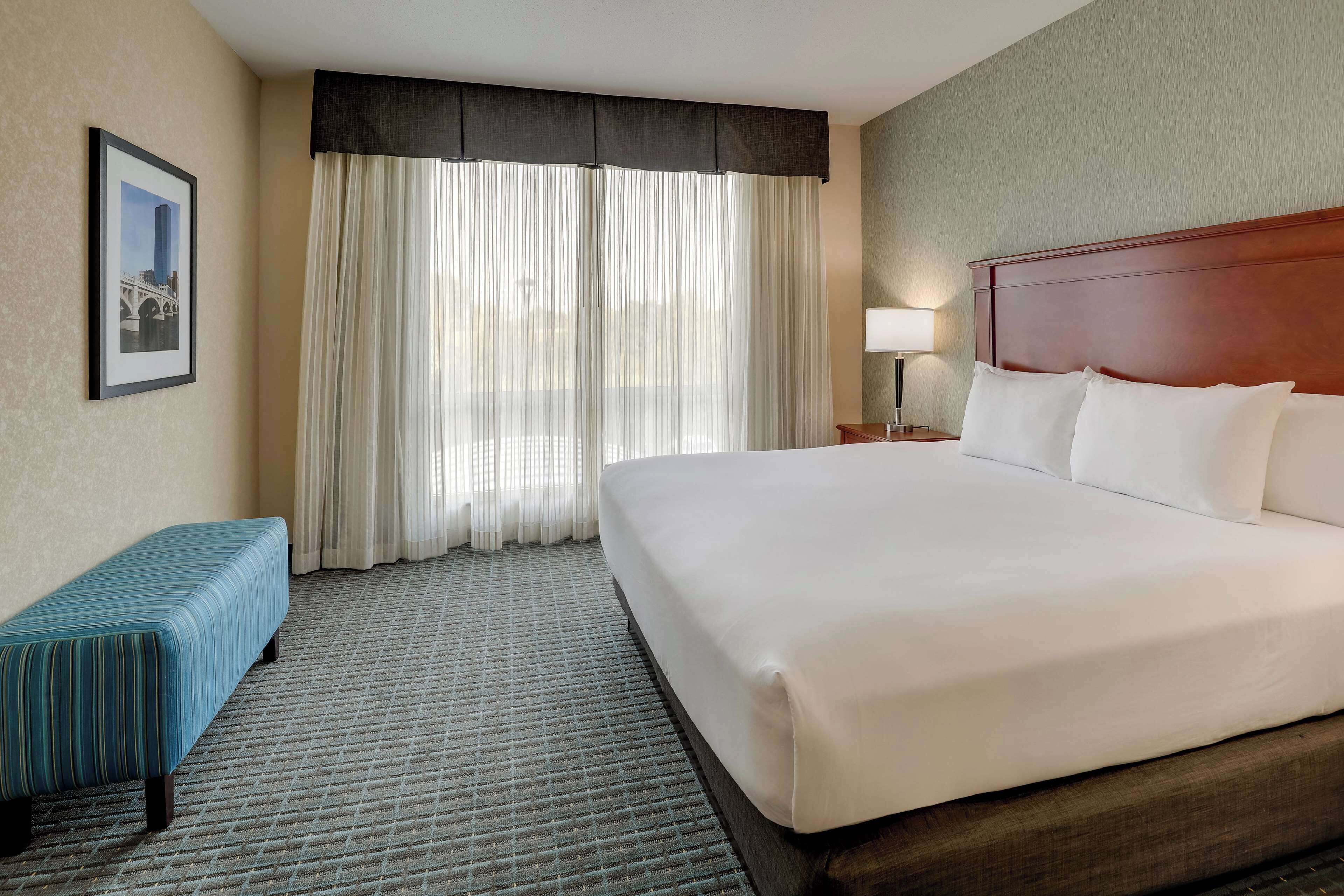 A hotel room with a large bed, a bench, a framed picture on the wall, and a window with curtains.