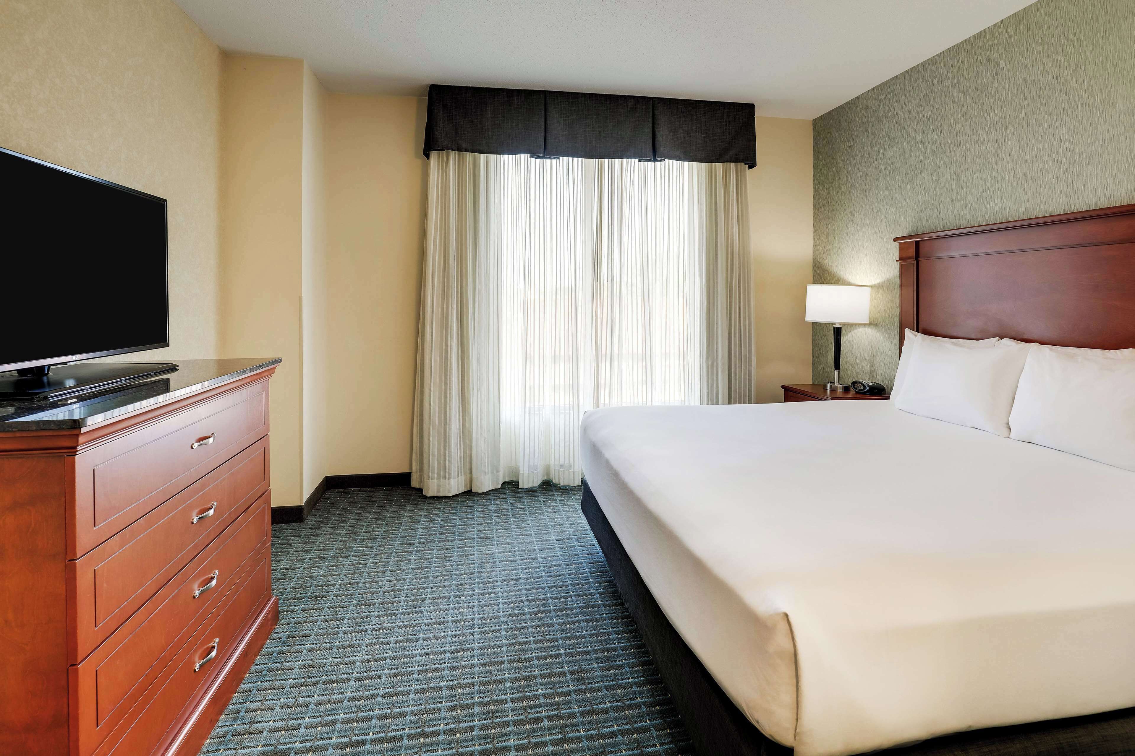 A hotel room with a large bed, a TV on a wooden dresser, a nightstand with a lamp, and a window with sheer curtains.