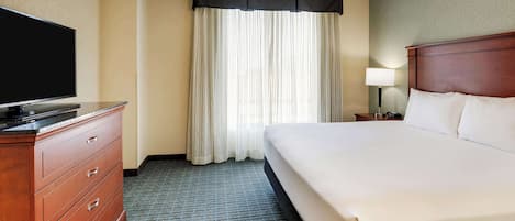 A hotel room with a large bed, a TV on a wooden dresser, a nightstand with a lamp, and a window with sheer curtains.