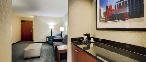 A hotel room with a kitchenette, a sofa, a small table, and a framed artwork on the wall.