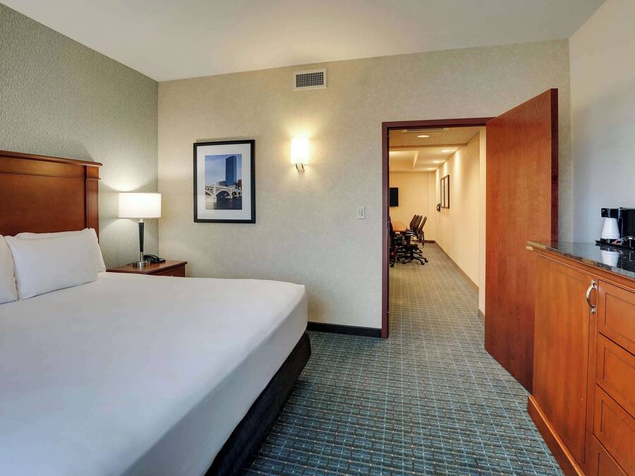 A hotel room with a large bed, a nightstand, a desk with a lamp, a framed picture on the wall, and a wooden cabinet.