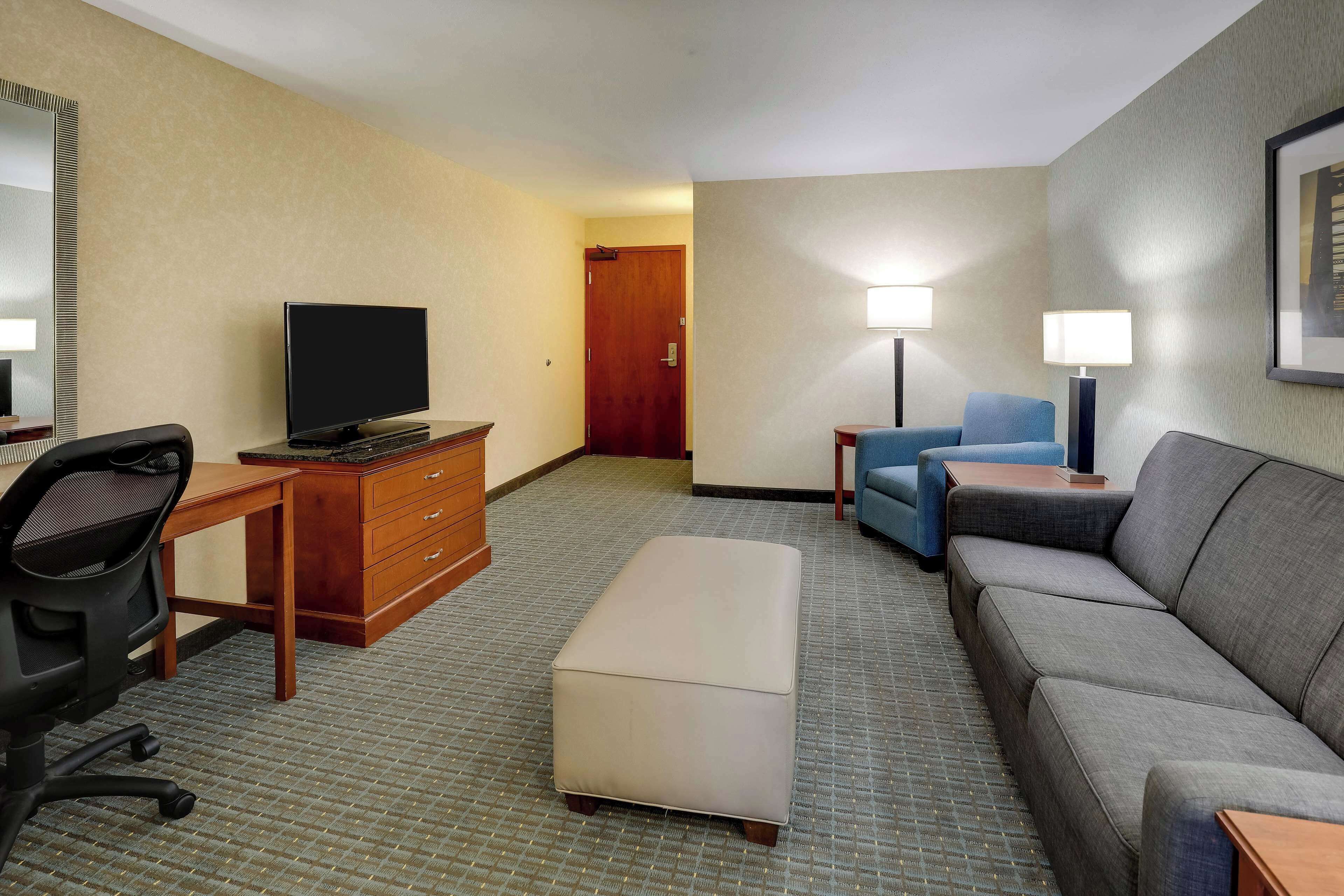 A hotel room with a desk, a flat-screen TV, a sofa, an ottoman, and two bedside lamps.