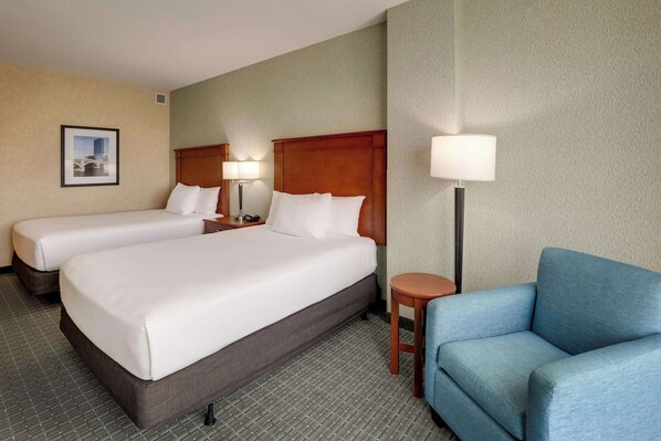 A hotel room with two beds, a blue armchair, a small table, and a lamp.