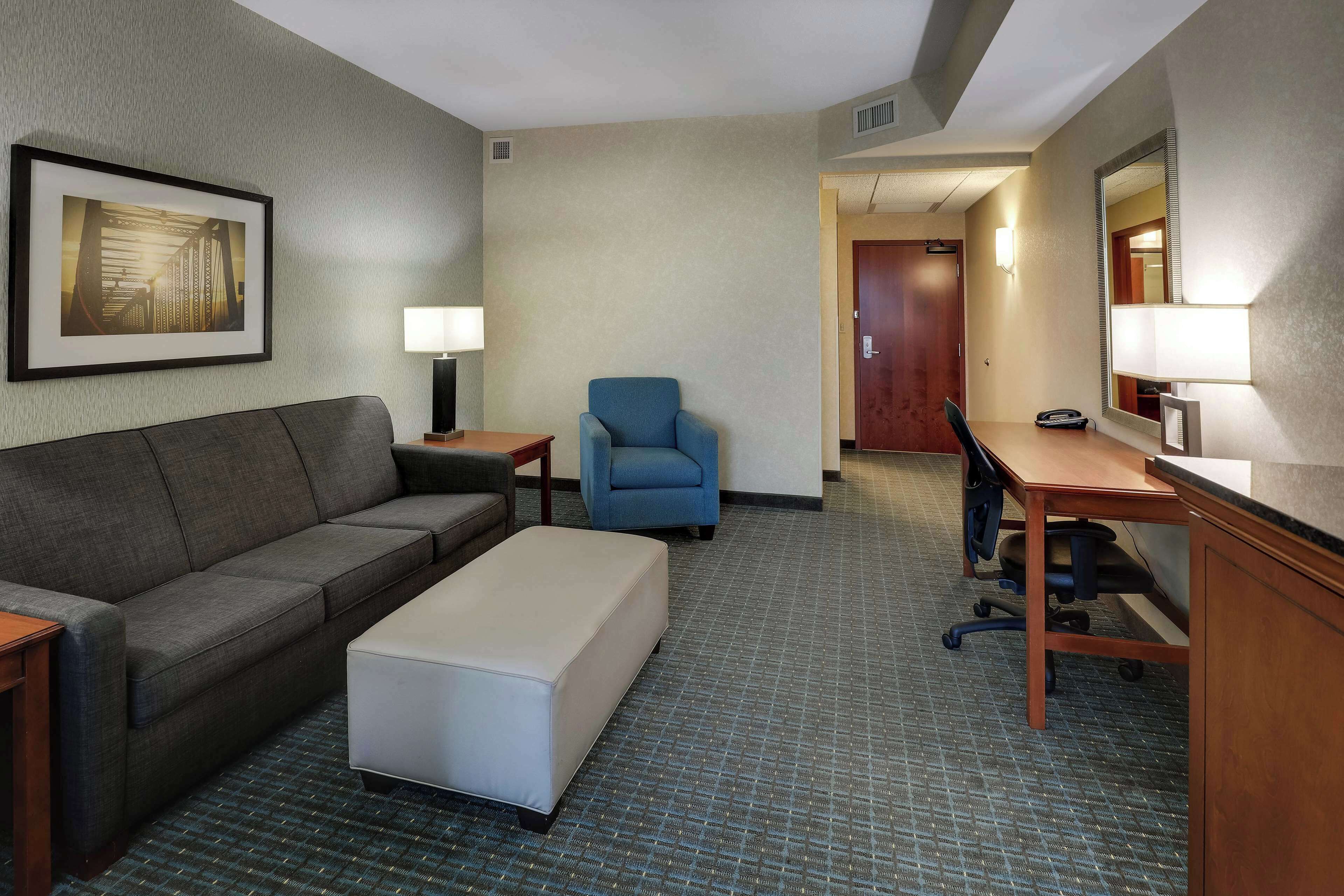 A hotel room with a gray sofa, a blue armchair, a desk with a chair, a small table, and a lamp.