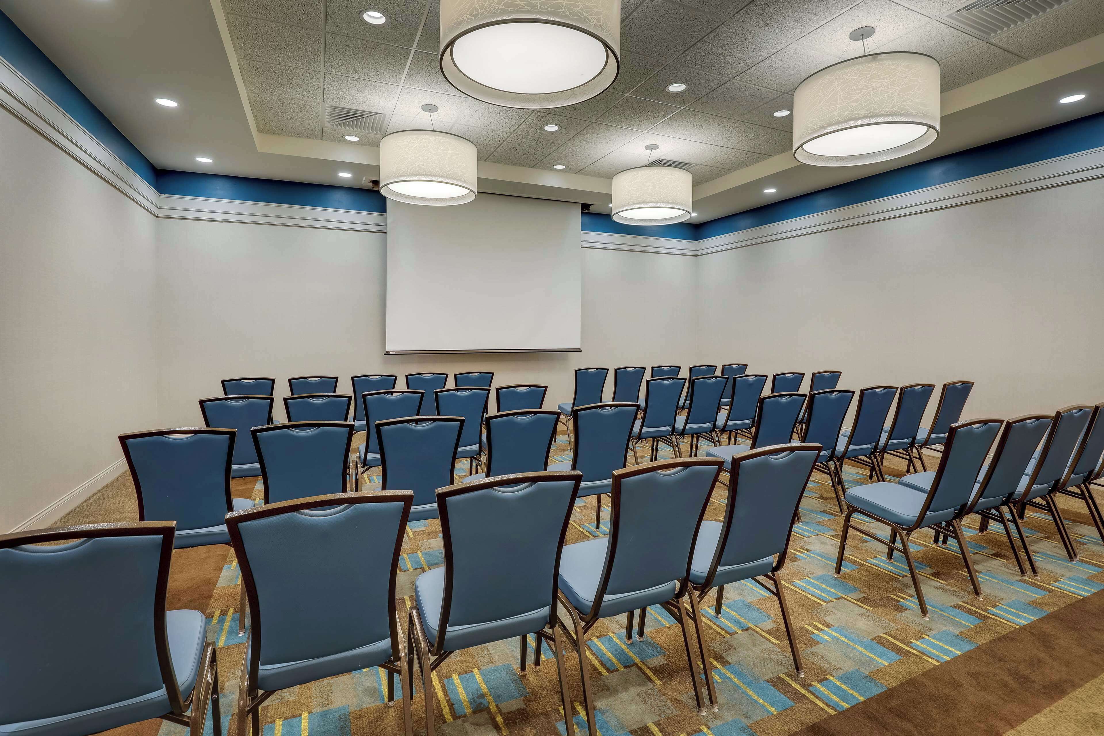 A conference room with rows of blue chairs, a large projection screen, and overhead lighting.