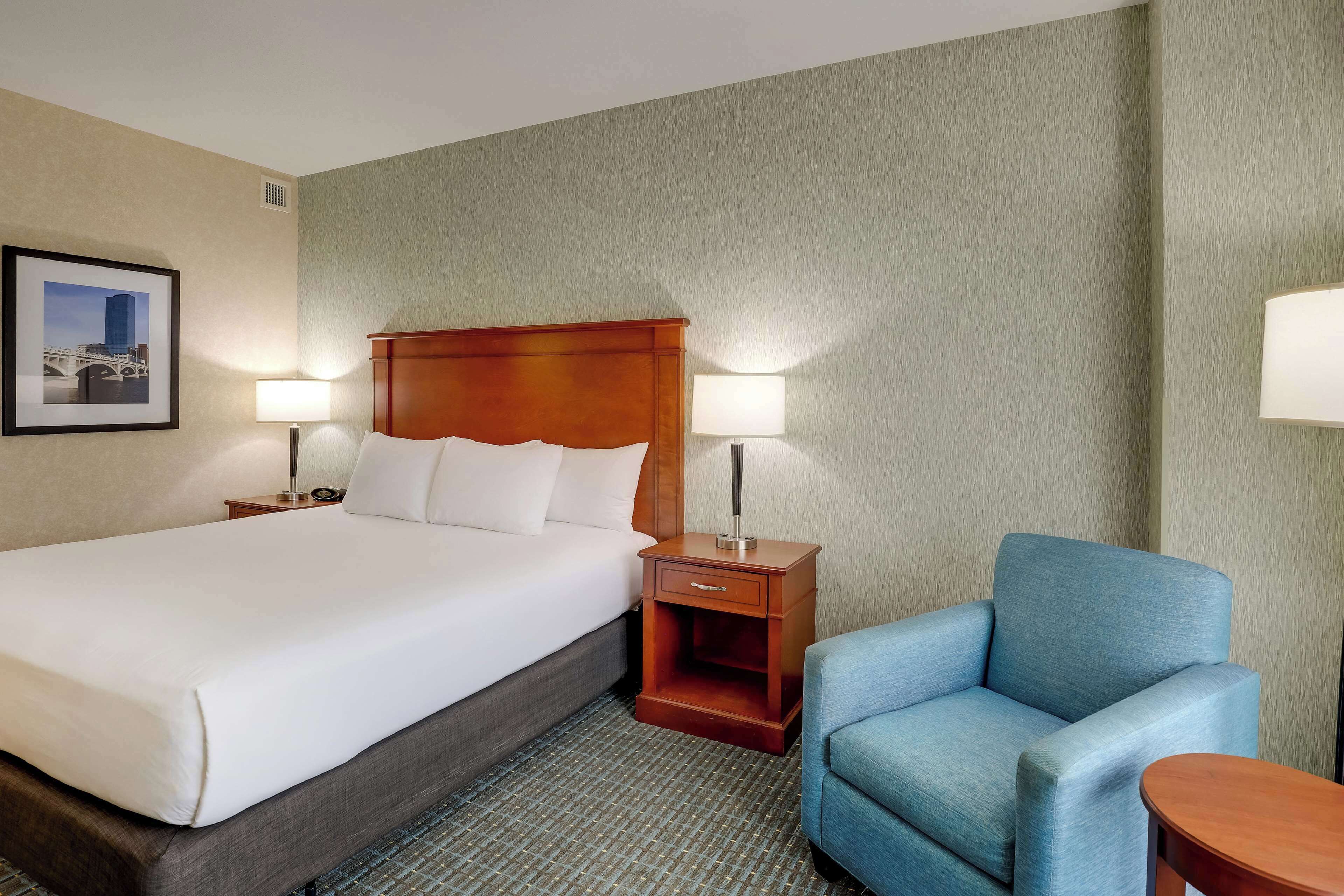 A hotel room with a large bed, two bedside tables with lamps, a blue armchair, and a framed picture on the wall.