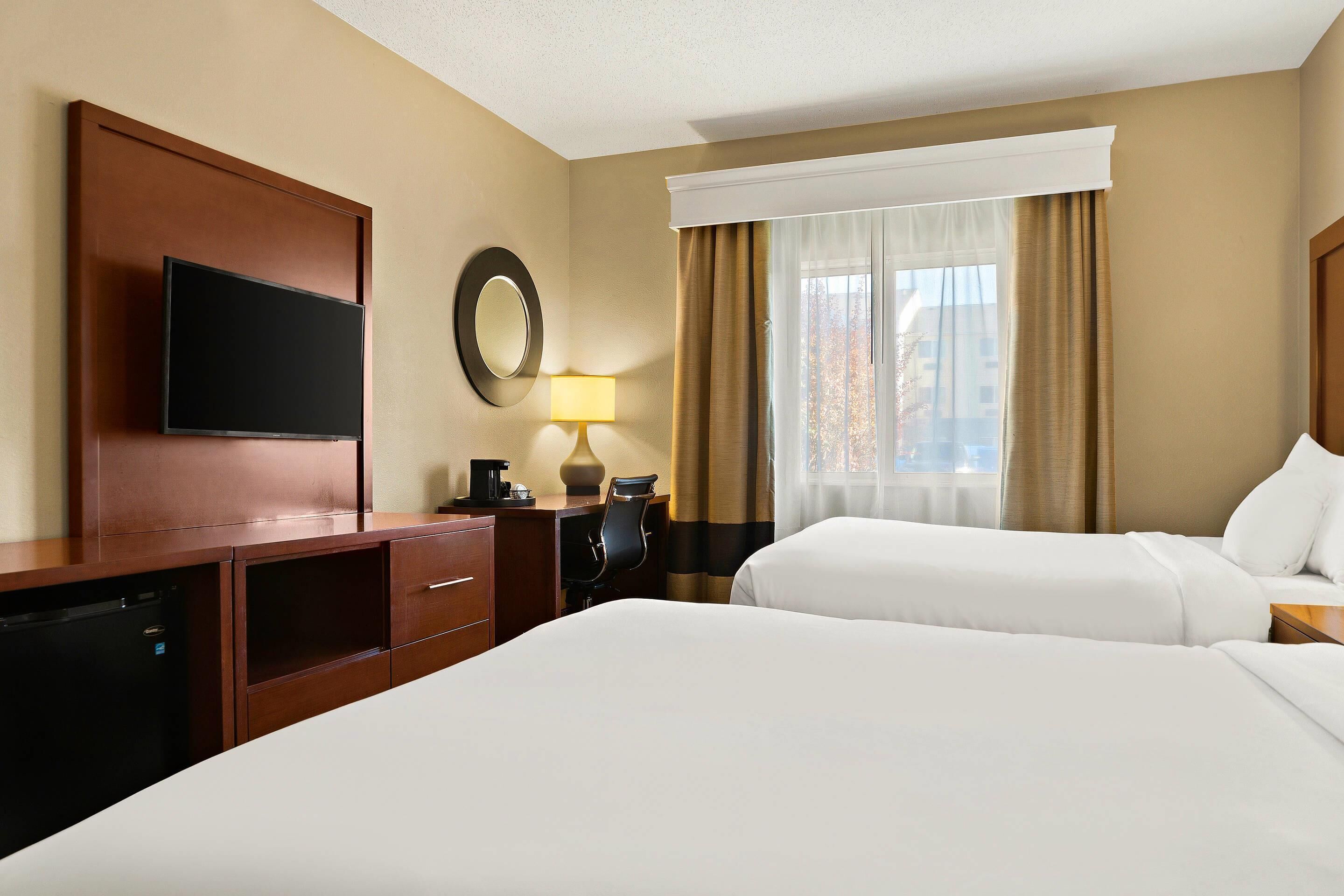 A hotel room with two beds, a TV, a desk with a lamp, and a window with curtains.
