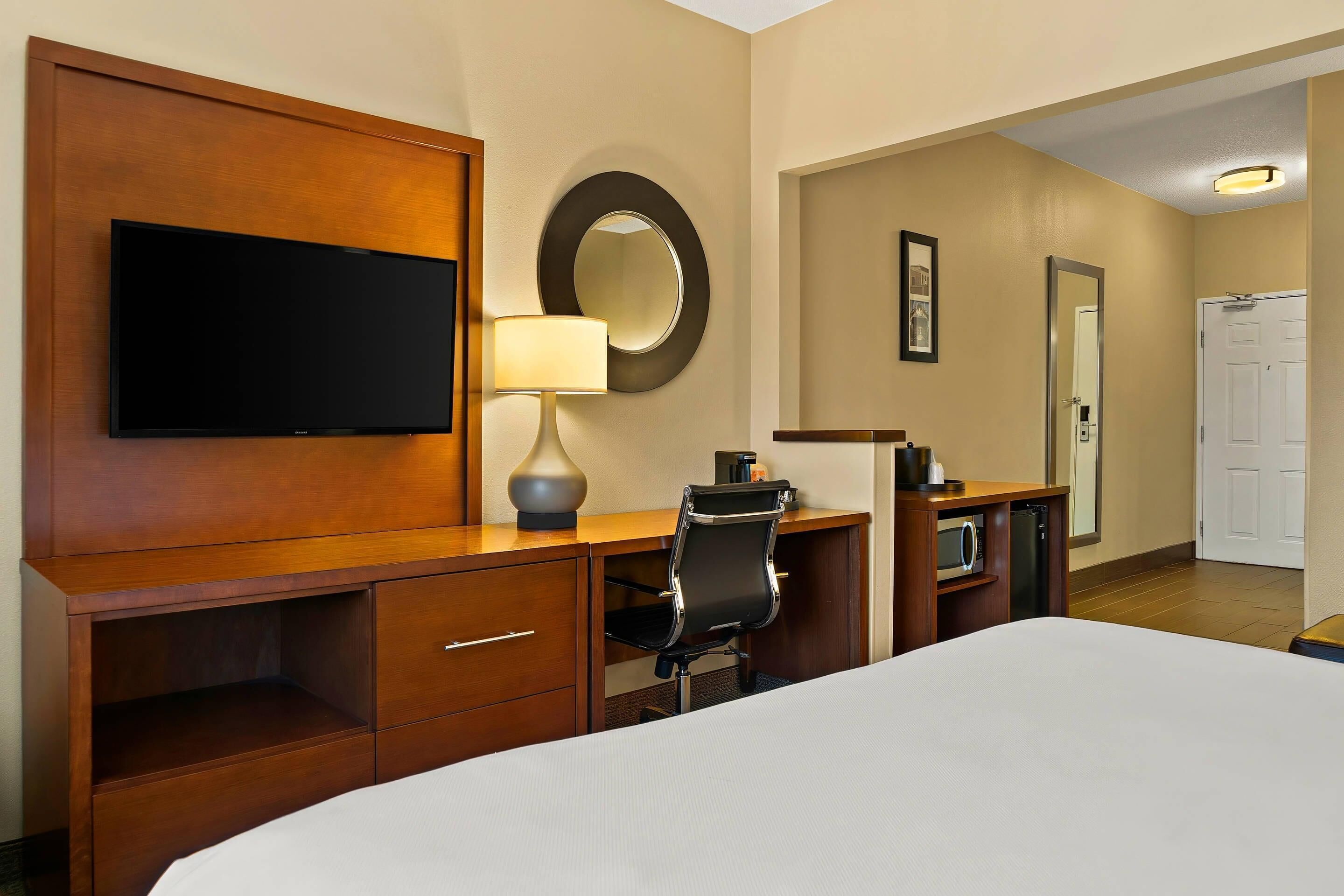 A hotel room with a bed, a desk with a chair, a television, a lamp, a mirror, and a door.