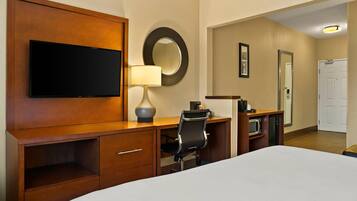 A hotel room with a bed, a desk with a chair, a television, a lamp, a mirror, and a door.