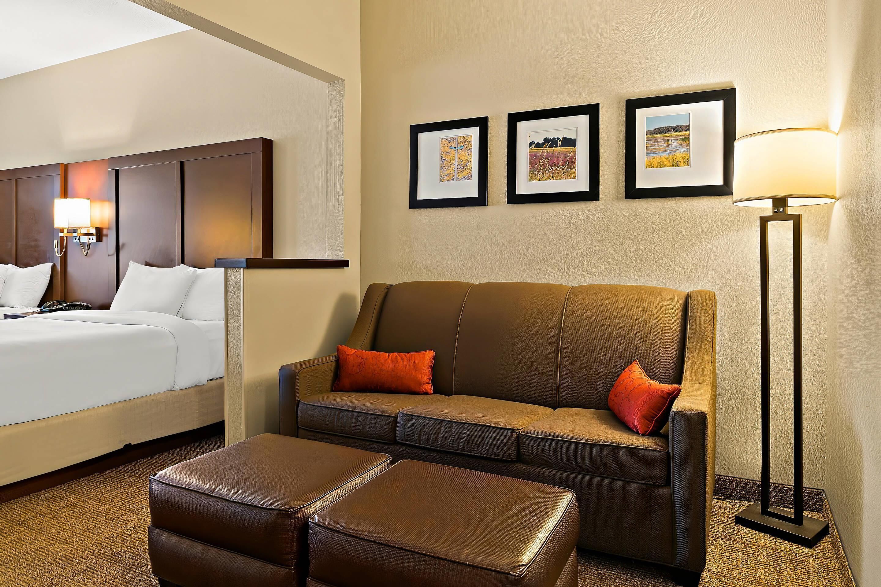 Una habitación de hotel con una cama, un sofá, un reposapiés y una lámpara de pie. Hay tres cuadros en la pared.