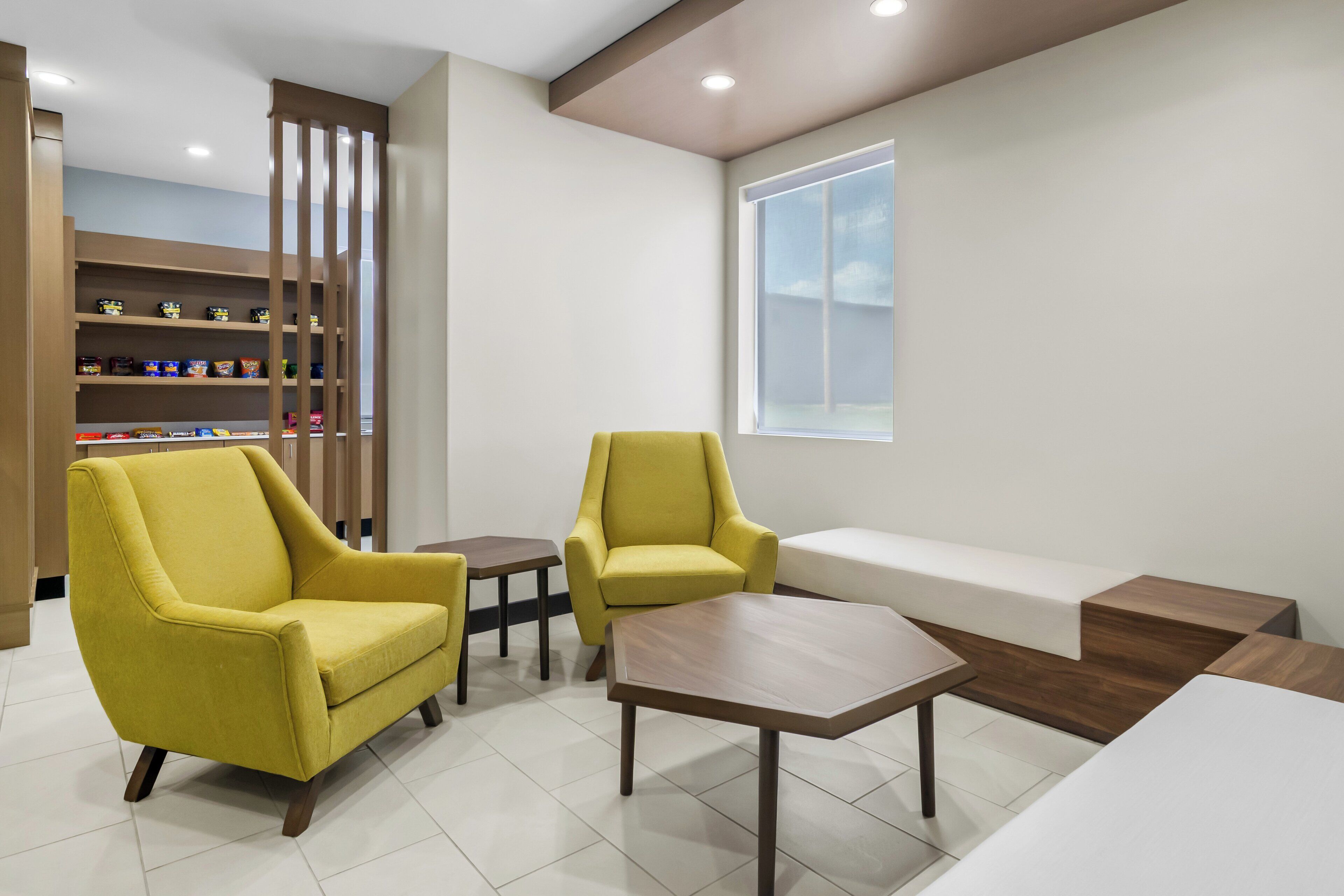 A modern waiting area with yellow armchairs, a wooden coffee table, and a bench. Shelves with snacks are in the background.