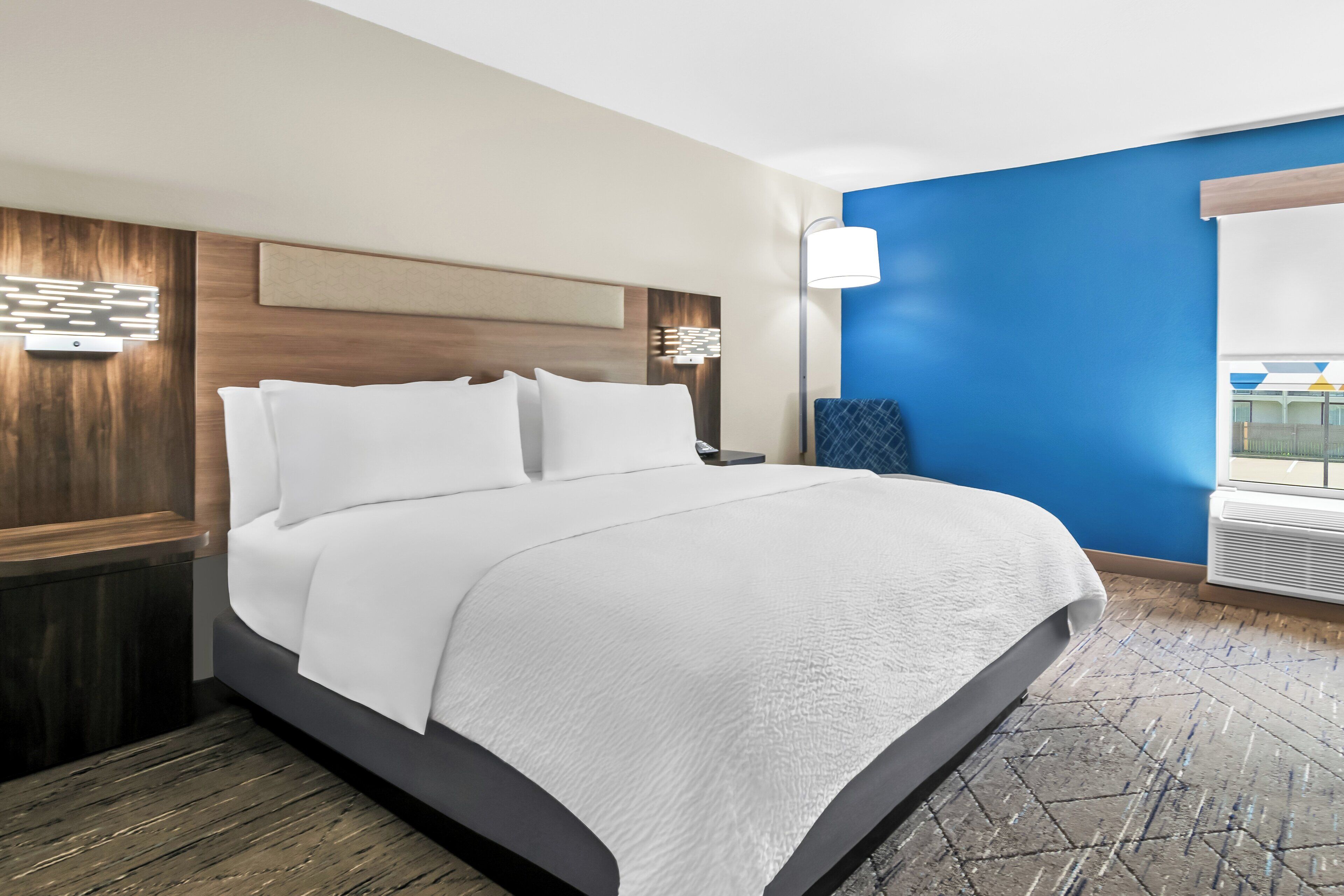 A hotel room with a large bed, a nightstand, a wall-mounted lamp, and a window with a view.