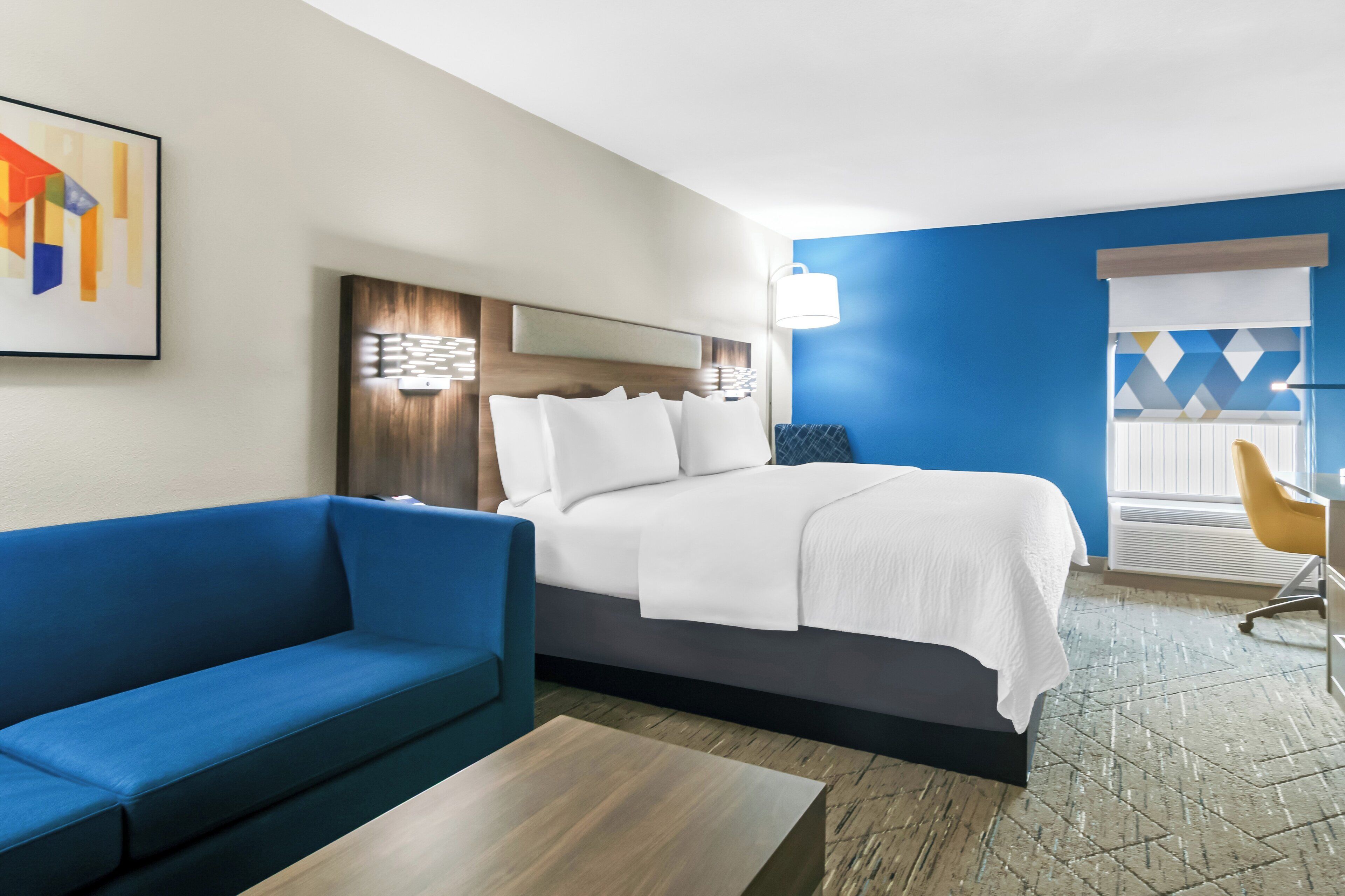 A hotel room with a bed, a blue sofa, a wooden coffee table, and a desk with a chair.