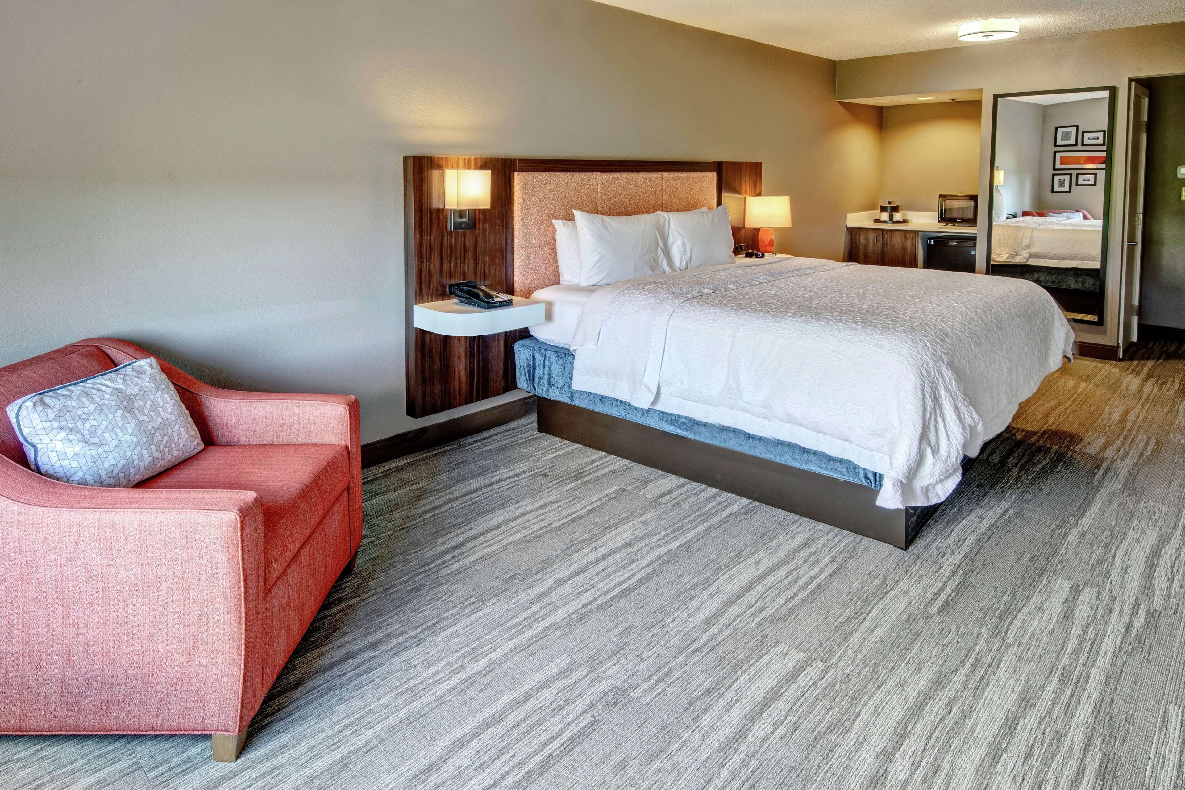 A hotel room with a large bed, a red armchair, a nightstand with a lamp, and a desk with a computer.