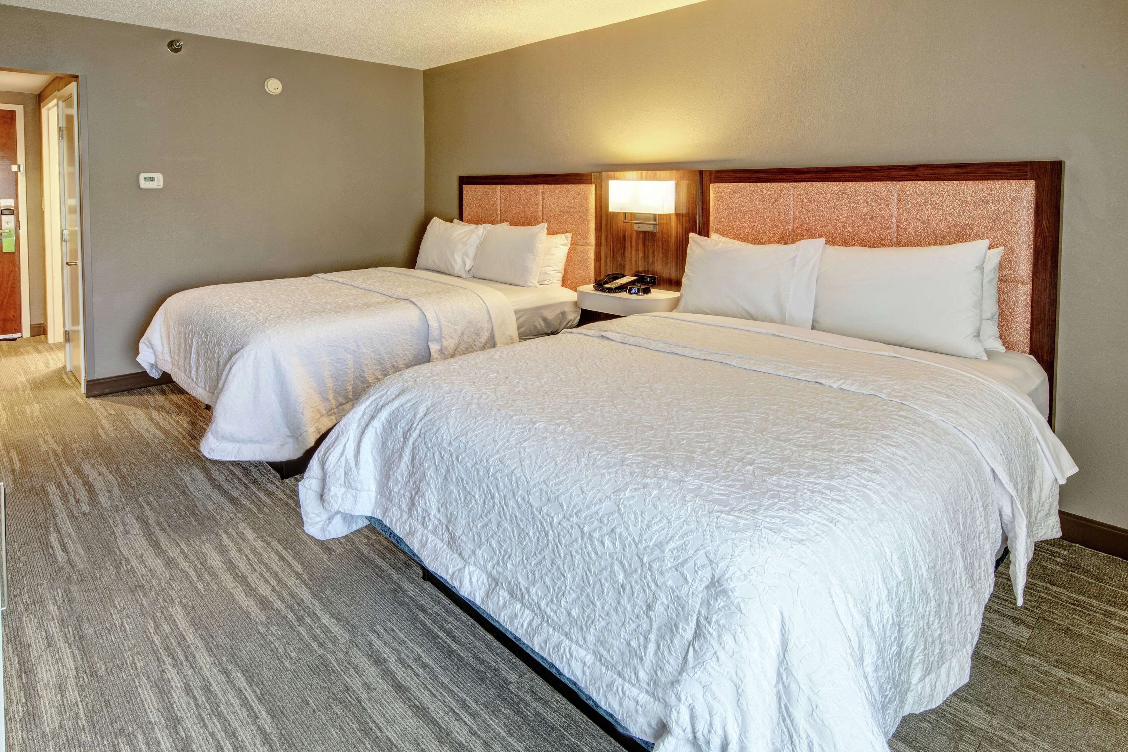 A hotel room with two beds, a nightstand with a lamp, and a door leading to another room.