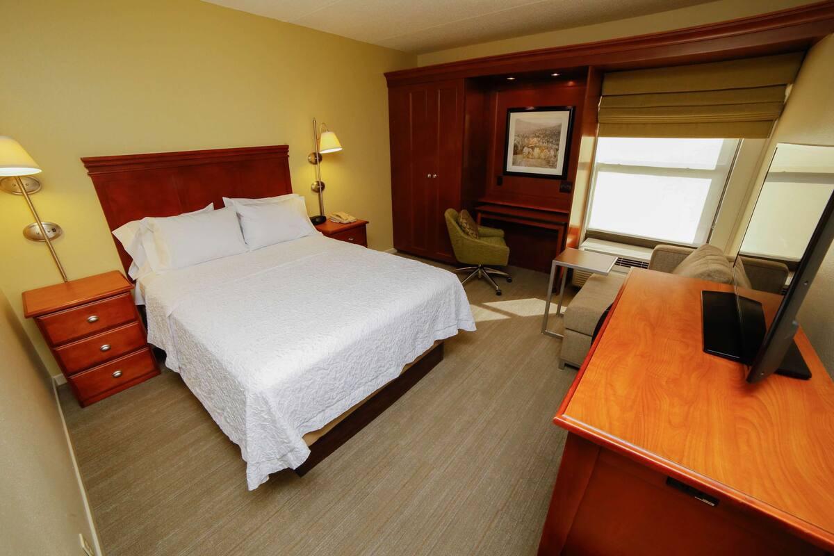 A hotel room with a large bed, a desk with a computer, a chair, a desk lamp, and a window with a view.