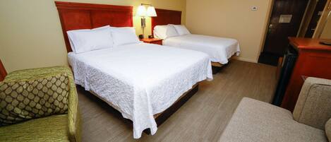 A hotel room with two beds, a nightstand with a lamp, a green patterned armchair, and a beige armchair.