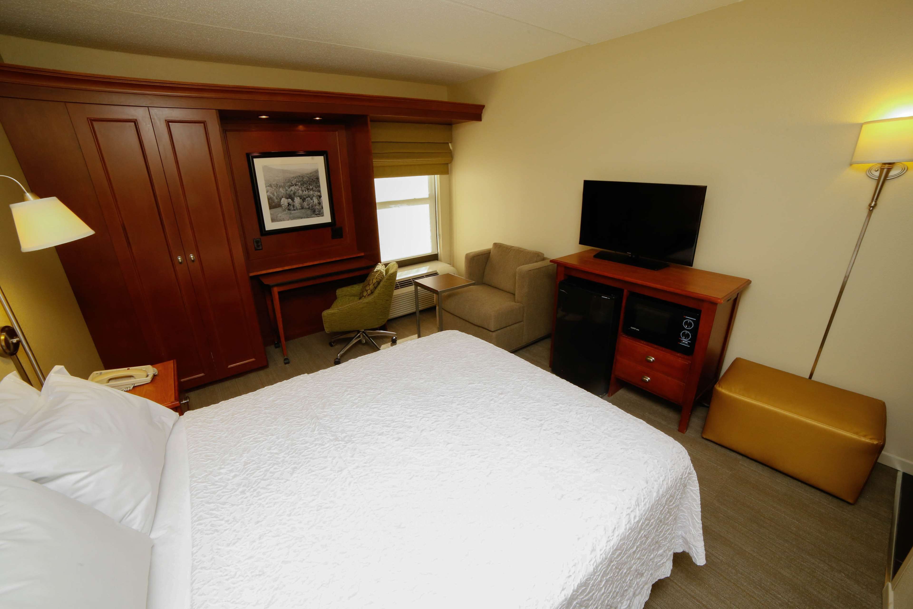 A hotel room with a large bed, a desk with a television, a chair, a desk lamp, and a wardrobe.