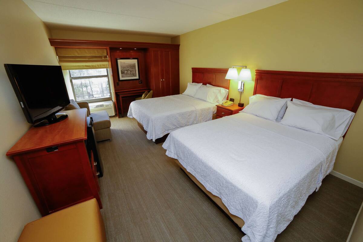 A hotel room with two beds, a television, a desk, and a window with blinds.