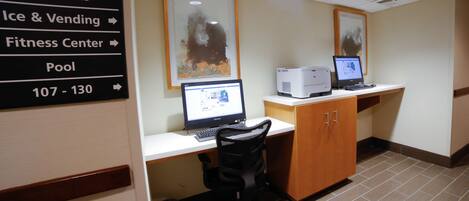 A hotel room with a desk, two computers, a trash bin, and a sign indicating amenities like meeting rooms, restrooms, ice & vending, a fitness center, and a pool.