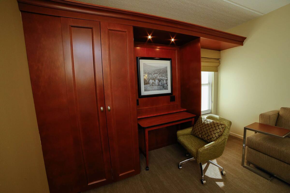 A room with a wooden wardrobe, a desk with a chair, and a framed picture on the wall.