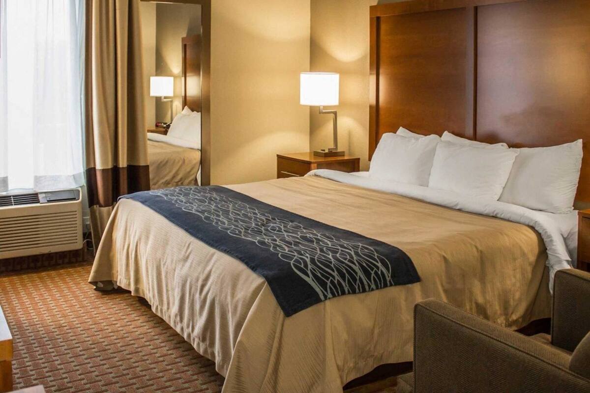 A hotel room with a large bed, a bedside table, a lamp, a chair, and a window with curtains.