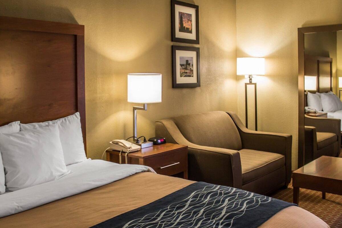 A hotel room with a bed, a chair, a desk, a nightstand with a phone, and two wall-mounted lamps.